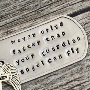 May include: A silver metal tag with the words "Never drive faster than your guardian angel can fly" embossed on it. The tag is attached to a silver angel wing charm.