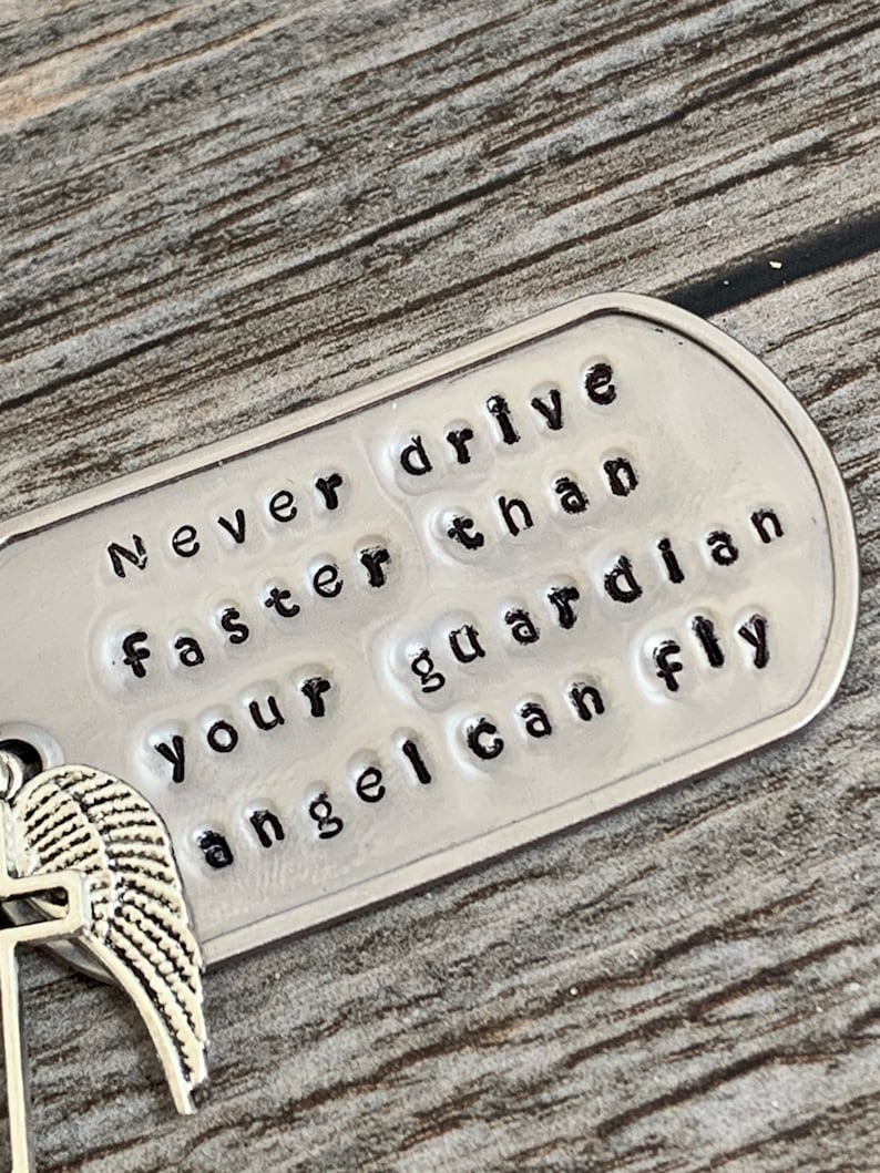 May include: A silver metal tag with the words "Never drive faster than your guardian angel can fly" embossed on it. The tag is attached to a silver angel wing charm.