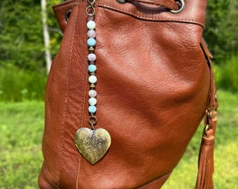 May include: A brown leather bucket bag with a drawstring closure and a decorative tassel. A beaded charm with a heart-shaped locket hangs from the bag. The charm features a mix of colorful beads.