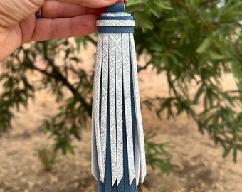 May include: A blue and white leather tassel keychain with a gold-colored clasp. The tassel features a geometric pattern and is made of layered leather strips. The keychain is designed for attaching to keys or bags.