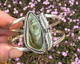 May include: A silver cuff bracelet featuring a green and brown stone set in a silver bezel. The stone is surrounded by two silver feathers.