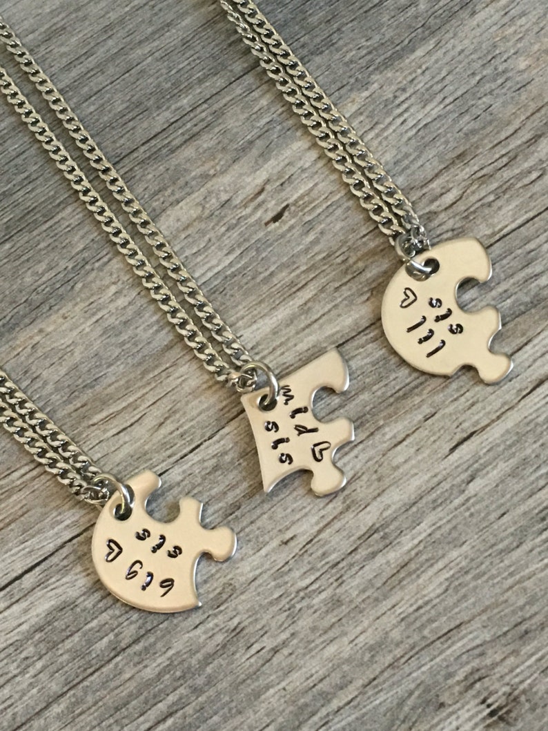 May include: Three silver necklaces with puzzle piece pendants. The pendants say "Big Sis", "Lil Sis", and "My Lil Sis".