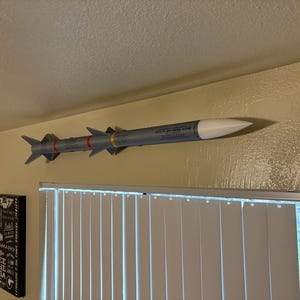 May include: A gray and white model missile is mounted on a wall. The missile has red and yellow bands, with text printed on the side. The missile is above a window with white blinds.