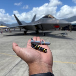 Könnte beinhalten: Eine Hand, die zwei Miniaturraketen hält, eine graue mit roten Akzenten und die andere schwarz mit gelben Akzenten. Im Hintergrund befindet sich ein graues Kampfflugzeug unter einem bewölkten blauen Himmel.