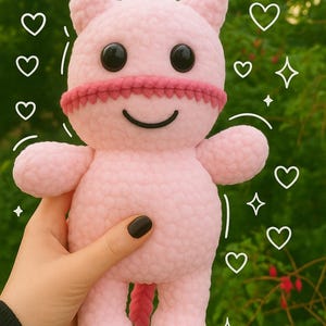 May include: A handmade pink plush toy shaped like a cat with black button eyes, a pink mouth, and a red band around its face. The toy has small ears and a tail. The background is a blurred green with drawn hearts and stars.