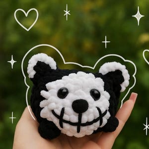 Könnte beinhalten: Ein handgefertigter schwarz-weißer Häkelbär mit großen schwarzen Augen und einem aufgestickten Lächeln. Der Bär hat abgerundete Ohren und ein flauschiges weißes Gesicht und eine weiße Brust. Das Spielzeug wird in einer Hand vor einem verschwommenen grünen Hintergrund mit weißen Stern- und Herz-Doodles gehalten.