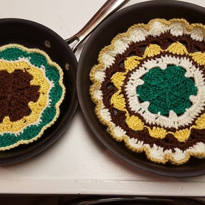 May include: Two crocheted sunflower pot holders in brown, white, yellow, and green colors. The pot holders are placed inside two black frying pans.