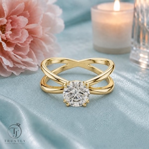 May include: A gold ring featuring a round brilliant-cut clear gemstone set in a four-prong setting. The band is designed with two crossing, polished gold strands that meet at the center stone, creating an open, X-shaped silhouette.