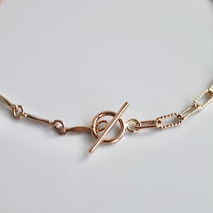 May include: A rose gold-toned chain necklace featuring a toggle clasp closure. The necklace is composed of alternating link styles, including smooth elongated bars and textured rectangular links, connected by small circular jump rings.