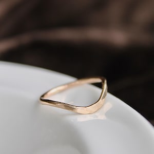 Puede incluir: Un anillo dorado con un diseño curvo y martillado. El anillo se muestra sobre una superficie blanca, con un fondo marrón borroso. La forma y textura únicas del anillo se destacan en esta toma de cerca.