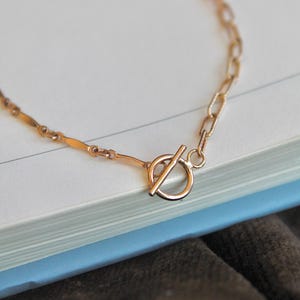 Könnte beinhalten: Eine goldfarbene Halskette mit Knebelverschluss. Die Kette weist eine Mischung aus länglichen und rechteckigen Gliedern auf. Die Halskette liegt auf einem Buch mit blau-weißem Einband, im Hintergrund befindet sich ein brauner Stoff.