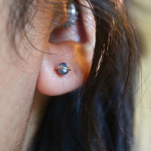 May include: A close-up of a person's ear with a small, gold-colored stud earring. The earring features a faceted, blue-grey gemstone encircled by a thin gold wire. Dark hair frames the ear.