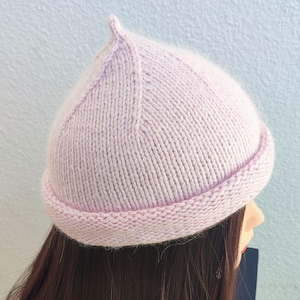 May include: A light pink knitted beanie hat with a rounded crown and a small, pointed top. The hat has a folded brim and is made of a soft, textured yarn. The hat is designed to be worn on the head.