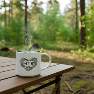May include: A white coffee mug with a green rim and a heart-shaped design made of gears and the letters "MTB" is on a wooden table. Steam rises from the coffee, suggesting it is hot. The background is a blurred forest scene.