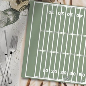 Può includere: Una tovaglietta rettangolare verde salvia con una stampa grafica bianca che riproduce un campo da football americano. Il design include linee delle yard, segni di riferimento e i numeri 10, 20, 30, 40 e 50 orientati in due direzioni.