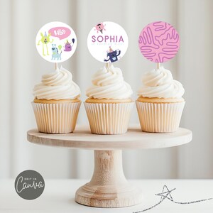 May include: Three cupcakes with white frosting topped with round monster-themed cupcake toppers. The toppers feature a purple monster with a party hat, a pink and purple abstract design, and a pink and purple monster with the text "Sophia is turning five".