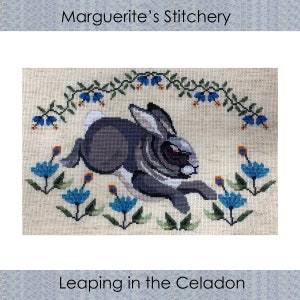 May include: Cross-stitch artwork featuring a leaping rabbit in shades of gray and white, surrounded by blue flowers and green foliage. The text "Leaping in the Celadon" is at the bottom, with "Marguerite's Stitchery" at the top.