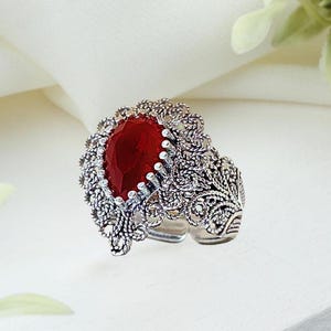 May include: A silver ring with an oval, deep red gemstone. The ring features an ornate, filigree design around the stone and along the band. The ring is set against a white background with floral accents.