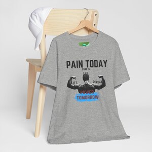 May include: A gray t-shirt with the text "PAIN TODAY" above a graphic of a muscular person flexing. Below the graphic are the words "GRIND POWER TOMORROW" in blue and red. A white baseball cap rests on a wooden chair.