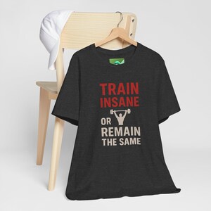 May include: Dark gray t-shirt with the text "TRAIN INSANE OR REMAIN THE SAME" in red and white. The shirt features a weightlifting graphic. A white baseball cap rests on a wooden chair.