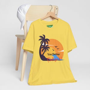 May include: Yellow t-shirt with a graphic of a sunset, palm trees, and the text "You Better Belize!" A white baseball cap rests on the back of a wooden chair. The t-shirt is hanging on a wooden hanger.