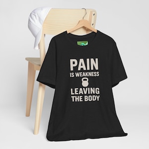 May include: Black t-shirt with the text "PAIN IS WEAKNESS LEAVING THE BODY" in white, along with a kettlebell graphic. A white baseball cap hangs on a wooden chair in the background.