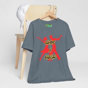 May include: A heather gray t-shirt with a red graphic of dancing figures and the words "Vamos a Bailar" in yellow. The shirt is hanging on a wooden hanger, with a white baseball cap resting on the chair behind it.