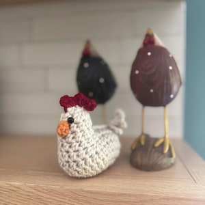 May include: A crocheted beige chicken figurine with a red comb and orange beak sits on a wooden surface. Two decorative chicken figures with brown and black bodies and white polka dots are in the background. The figurines are a farmhouse style.