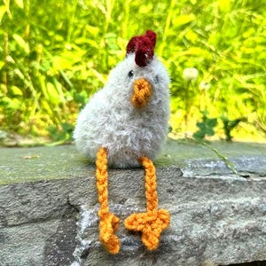 May include: A handmade, fluffy white chicken figurine with a red comb and orange beak and feet. The chicken is sitting on a stone surface with a green background. The chicken's feet are dangling.