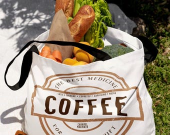 May include: White tote bag with black straps, filled with groceries, including bread, carrots, and broccoli. The bag features a brown circular design with the word "COFFEE" and the text "THE BEST MEDICINE FOR A 12-HOUR SHIFT". Brown sunglasses rest on a white blanket.