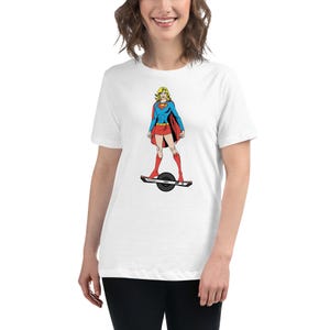 Puede incluir: Camiseta blanca con una ilustración de estilo cómic de una superheroína con top azul, falda roja y capa, de pie sobre un monopatín eléctrico de una rueda. La superheroína tiene el pelo rubio y botas rojas.