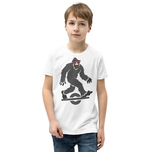 May include: White t-shirt featuring a gray Bigfoot character wearing pink sunglasses and riding a one-wheel board. The Bigfoot has a brown mustache. The shirt is worn by a person in blue jeans.