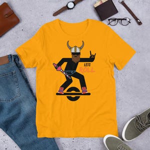 May include: Mustard yellow t-shirt featuring a cartoon Viking riding a one-wheel electric skateboard. The Viking has a brown beard, a horned helmet, and is holding a pink and silver weapon. The text "Lets Roll" is printed on the shirt.