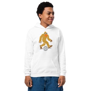 May include: White hoodie featuring a Bigfoot graphic riding a one-wheel electric skateboard. The Bigfoot is depicted in shades of brown, wearing sunglasses and a red beak. The hoodie has a front pocket and a drawstring hood.