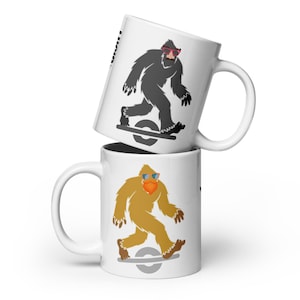 May include: Two white ceramic mugs stacked on top of each other. Each mug features a cartoon Bigfoot character riding a one-wheel electric skateboard. One Bigfoot is gray with red sunglasses, and the other is tan with blue sunglasses.