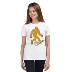 May include: White t-shirt featuring a cartoon Bigfoot character wearing sunglasses and riding a one-wheel electric skateboard. The Bigfoot is brown with a heart-shaped orange chest. The skateboard is gray.