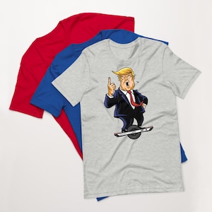 May include: Three short-sleeved t-shirts in red, blue, and grey. The grey shirt has a cartoon illustration of a man in a suit riding a one-wheel device, pointing upwards with an open mouth.