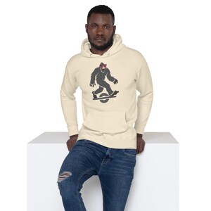 May include: Cream-colored hoodie with a Bigfoot graphic riding a one-wheel electric skateboard. The Bigfoot is depicted in gray with a red bandana. The hoodie has a front pocket and a drawstring hood. The person is wearing blue jeans.
