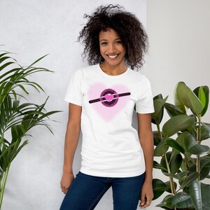 May include: White t-shirt featuring a pink heart design with a black and pink graphic. The graphic includes a circle and a horizontal line. The shirt is made of a soft material and is suitable for casual wear.