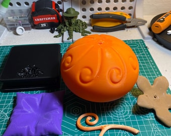 May include: A 3D printed orange sphere with a swirling pattern, a separate tan stem piece, a small purple square cushion, and a tan star-shaped base. These items are arranged on a green cutting mat alongside a black box and various tools.