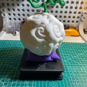 Puede incluir: Una escultura blanca en forma de nube con una parte superior verde en forma de vid, que descansa sobre una pequeña almohada morada y una base negra. La escultura tiene una superficie detallada y texturizada. La base parece un libro o una caja.