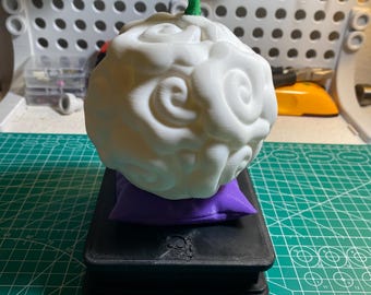 May include: A white, cloud-shaped sculpture with a green, vine-like top, resting on a small purple pillow and a black base. The sculpture has a detailed, textured surface. The base appears to be a book or box.