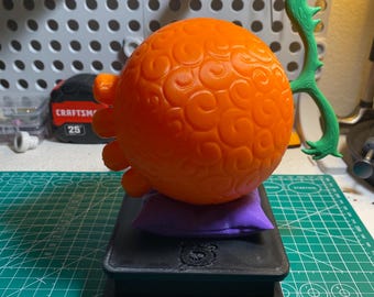 May include: An orange, spherical object with a textured surface and green, curved appendage. It rests on a small purple pillow atop a black, rectangular base. The base has a bear logo.