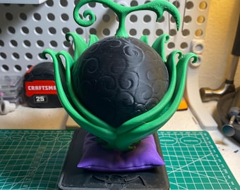 May include: A 3D-printed replica of a black Devil Fruit, with green leaves and stem, resting on a purple pillow and a black base. The fruit has a detailed, textured surface.