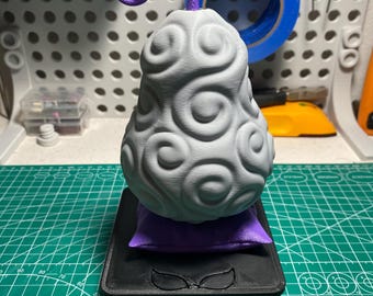 May include: A 3D-printed gray devil fruit with a purple stem, resting on a purple pillow and a black base. The fruit has a swirling pattern and is set against a white pegboard background.
