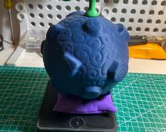 May include: A 3D-printed, dark blue fruit with a green stem, resting on a purple pillow and a black base. The fruit has a textured surface with swirling patterns. The base is rectangular with a gear-like design.