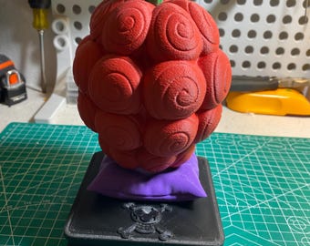May include: A red, spiral-textured fruit with green leaves, resting on a purple pillow and a black, skull-adorned box. The fruit is a vibrant red, and the leaves are a bright green. The box has a skull and crossbones design.