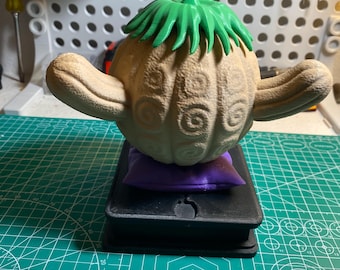 May include: A tan-colored, stylized fruit with a green leafy top and curved appendages. The fruit rests on a small purple pillow atop a black rectangular base. The design features swirling patterns.