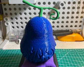 May include: A blue, pear-shaped sculpture with a green stem and curly tendril, resting on a purple pillow and a black base. The sculpture has detailed patterns and textures, resembling a stylized fruit.