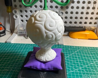 May include: A white, spherical fruit with a green stem and swirling patterns, resting on a purple pillow and a black base. The fruit is a 3D-printed model, likely a collectible or decorative item.
